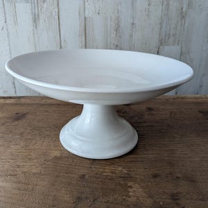 Puede incluir: Un cuenco de cerámica blanca sobre pedestal con un recipiente ancho y poco profundo y una base robusta y acampanada. La superficie lisa y brillante del plato presenta un borde redondeado limpio y un tallo central que sostiene el cuenco.