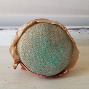 May include: A round, cream-colored pincushion with a green fabric center and a ruffled edge.