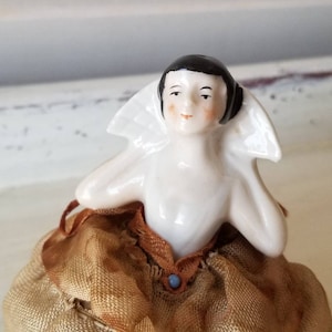 May include: A porcelain doll with black hair and a white collar sits on a brown fabric base. The doll has a painted face with rosy cheeks and a blue button on her dress.