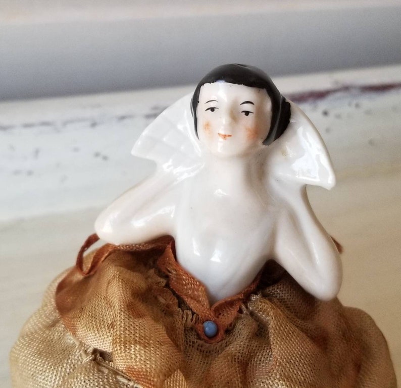 May include: A porcelain doll with black hair and a white collar sits on a brown fabric base. The doll has a painted face with rosy cheeks and a blue button on her dress.
