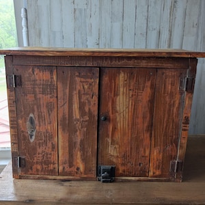 Può includere: Un mobile rustico in legno a due ante, con una calda finitura marrone-rossastra. Il mobile presenta venature del legno visibili e cerniere metalliche, suggerendo uno stile vintage o artigianale. È appoggiato su una superficie di legno.