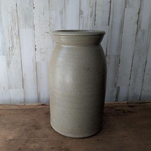 Op de afbeelding: Een cilindrische steengoed vaas met een gespikkelde lichtbeige afwerking en een brede, uitlopende rand. De vaas heeft een getextureerd, mat oppervlak over het hele lichaam, loopt iets taps toe naar de basis en heeft een neutrale kleur.