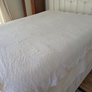 May include: A cream-colored, textured bedspread with intricate woven patterns. The bedspread is draped over a bed, showcasing its detailed design and soft, neutral color. The overall aesthetic is classic and inviting, suitable for a bedroom setting.