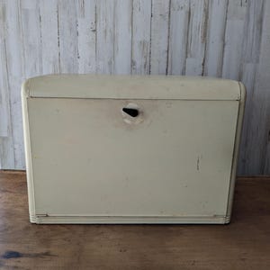 May include: A vintage cream-colored metal bread box with a rectangular shape and rounded top edges. The front panel features a central black handle mechanism with visible wear, scuffs, and minor surface discoloration across the exterior finish.