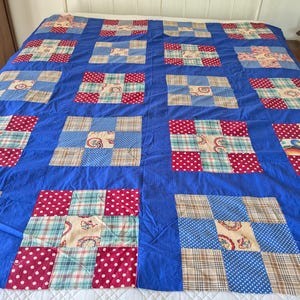 Może przedstawiać: Duża, ręcznie robiona kołdra z żywym niebieskim tłem. Kołdra ma wzór patchworkowy z kwadratami czerwonych kropek, kraty i wzorzystej tkaniny. Kołdra jest rozłożona na łóżku, prezentując swój rozmiar i skomplikowany design.