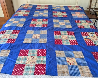 May include: A large, handmade quilt with a vibrant blue background. The quilt features a patchwork design with squares of red polka dots, plaid, and patterned fabric. The quilt is spread out on a bed, showcasing its size and intricate design.