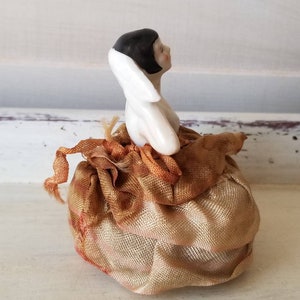 May include: A porcelain doll with black hair and a white dress sits on a gold satin cushion. The doll is looking to the right.