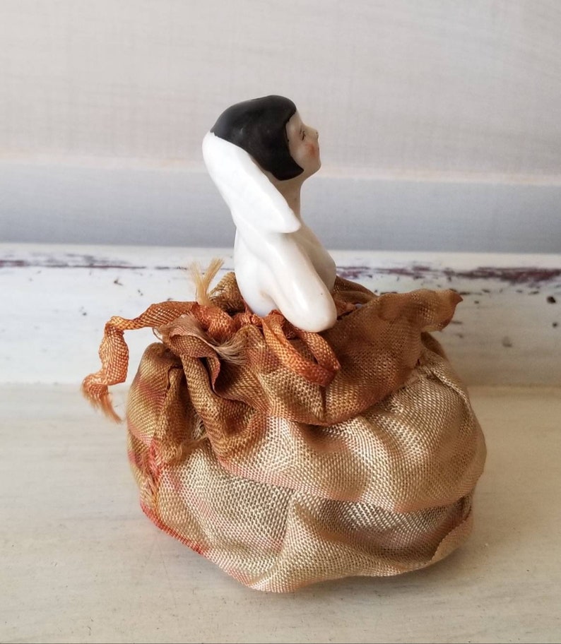 May include: A porcelain doll with black hair and a white dress sits on a gold satin cushion. The doll is looking to the right.