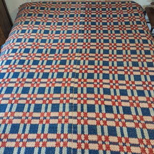 May include: A woven bedspread featuring a repeating geometric plaid pattern in shades of deep blue, bright red, and cream. The textile displays a structured grid design with small star-like motifs integrated into the intersections of the colored lines.
