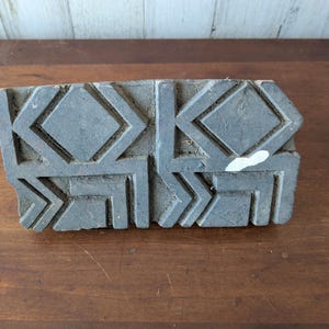 Puede incluir: Un bloque de madera gris desgastado con un patrón geométrico repetitivo de diamantes y formas angulares. El bloque es rectangular y parece tallado a mano, con textura visible y ligeras imperfecciones. Se ve una pequeña zona de pintura blanca.