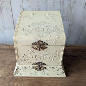 May include: Cream-colored vintage jewelry box with ornate floral and scrollwork detailing. Features two brass-toned clasps and decorative accents. The box has a hinged lid and a rectangular base, showcasing a classic, antique aesthetic.