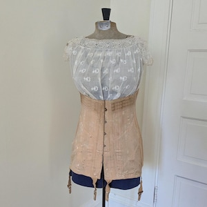 May include: A vintage-style beige corset with a front hook-and-eye closure and attached garter straps, worn over a white, sheer, short-sleeved blouse featuring delicate floral embroidery and a wide, intricate lace trim along the neckline.