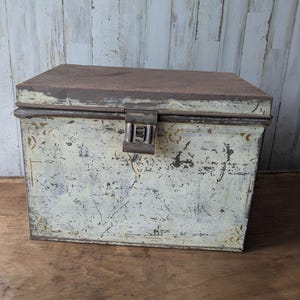 Può includere: Una scatola rettangolare in metallo con coperchio incernierato e chiusura centrale. L'esterno presenta una finitura color crema usurata con ruggine marrone scuro visibile, graffi e tenui motivi decorativi color oro lungo i bordi.