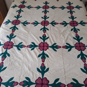 May include: A cream-coloured quilt featuring a repeating floral design. The flowers are a deep magenta, with teal green leaves and stems. The quilt is displayed on a bed, highlighting the detailed pattern and craftsmanship.