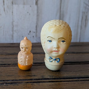 May include: Two vintage ceramic dolls. The larger doll is a girl with a white dress and a blue bow. The smaller doll is a boy wearing a brown and white uniform. Both dolls have painted faces and are in good condition.