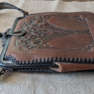 May include: Brown leather purse with a decorative metal clasp and a stitched leather strap. The purse features an embossed design on the front and is likely vintage. The strap is approximately 1 inch wide.