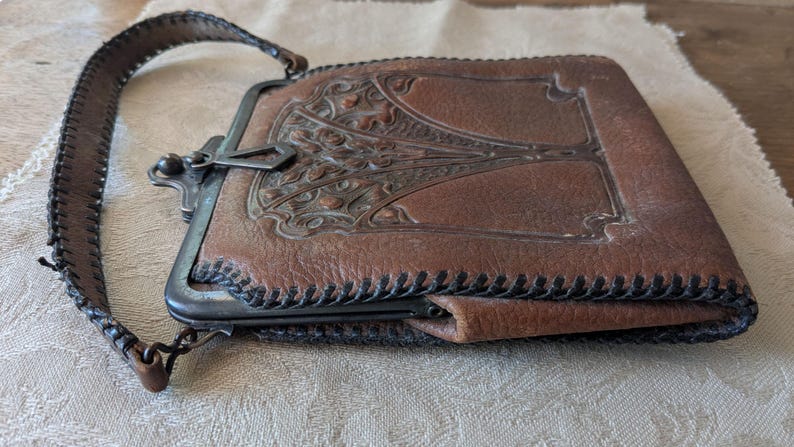 May include: Brown leather purse with a decorative metal clasp and a stitched leather strap. The purse features an embossed design on the front and is likely vintage. The strap is approximately 1 inch wide.