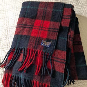 May include: A folded wool blanket featuring a dark navy blue and bright red plaid pattern with thin dark green accent lines. The blanket has a fringed edge and a small rectangular blue label with the text Pendleton sewn onto the fabric.
