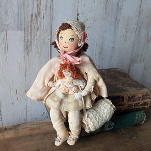 May include: A vintage-style cloth doll with brown hair, blue eyes, and a pink ribbon. The doll is wearing a light pink cape and holding a smaller doll. It is seated on a stack of books, with a white, textured bag nearby. The doll's dress is cream-coloured.