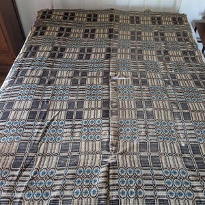 May include: A large rectangular woven coverlet featuring a repeating geometric pattern in shades of dark brown, cream, and muted blue. The textile displays a grid-like structure with alternating blocks of intricate woven motifs across the surface.