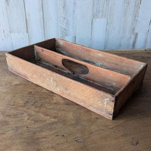 May include: A rectangular, vintage wooden tray with two compartments and a handle cutout. The tray has a weathered, aged appearance with visible wood grain and a warm, brown color. The tray is likely used for organizing or serving.