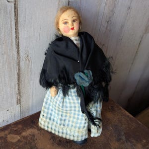 May include: A vintage doll with a painted face, wearing a black shawl with fringe, a blue and white checkered dress, and a blue flower accent. The doll has blonde hair and rosy cheeks, and is standing upright.