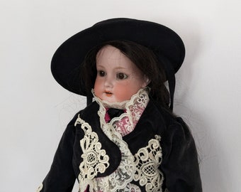 Puede incluir: Una muñeca de porcelana con cabello oscuro que lleva una chaqueta de terciopelo negro con ribetes de encaje blanco y un sombrero negro.