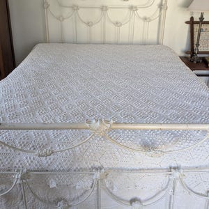 Pode incluir: Uma cama de metal branco com uma colcha branca estampada a condizer. A cama está numa divisão com chão de madeira e um armário de madeira. Uma pequena mesa com uma lâmpada e uma imagem emoldurada está ao lado da cama.