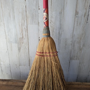 Könnte beinhalten: Ein Vintage-Besen mit rotem Stiel und einem Kopf aus Naturstroh. Der Stiel hat ein dekoratives Etikett in der Nähe der Oberseite. Der Strohkopf ist mit rotem und grauem Garn gebunden. Der Besen wird vor einem verwitterten Holzhintergrund gezeigt.