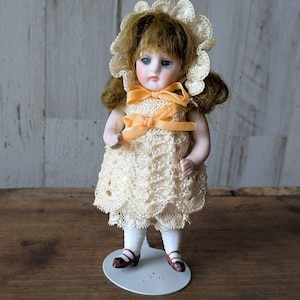 May include: A vintage porcelain doll with blue eyes, brown hair, and a cream-colored lace dress. The doll wears a matching bonnet and has a yellow ribbon. It stands on a metal stand, wearing white stockings and brown shoes.