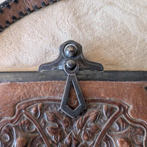 May include: Close-up of a brown leather purse with a decorative floral pattern. The purse has a metal clasp and a leather strap. The leather is textured and shows signs of wear, suggesting an antique or vintage item.