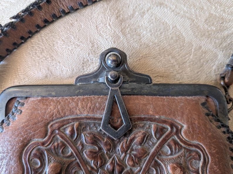 May include: Close-up of a brown leather purse with a decorative floral pattern. The purse has a metal clasp and a leather strap. The leather is textured and shows signs of wear, suggesting an antique or vintage item.