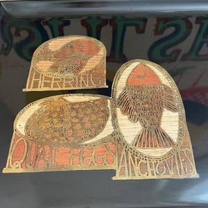 May include: A black rectangular serving tray with three decorative wooden inlays. The inlays feature fish and the words "HERRING", "QUAIL EGGS", and "ANCHOVIES" in gold lettering. The tray is on a wooden surface.