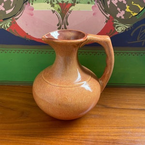 May include: A brown ceramic pitcher with a rounded body and a handle. The pitcher has a glazed finish and a subtle sheen.