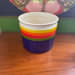 May include: A white ceramic utensil holder with a blue base and colorful stripes in yellow, orange, red, and green.