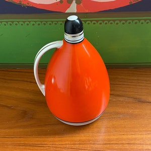 May include: Orange ceramic pitcher with a black and white lid and a gold trim. The pitcher has a handle and a rounded body.
