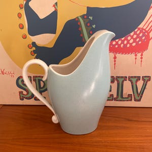 May include: A light blue ceramic creamer with a white handle and spout. The creamer has a speckled texture and a curved design. The background features a vintage poster with the word "SPELV".