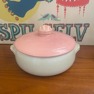 May include: A round ceramic casserole dish with a speckled off-white base and two side handles. The dish features a smooth, glossy pastel pink lid topped with a small, sculpted, flower-shaped handle in the same matching pink color.