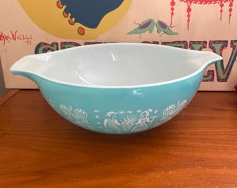 Peut inclure: Un bol mélangeur en verre turquoise et blanc avec deux poignées latérales. L'extérieur présente un motif graphique blanc répétitif composé de personnages, d'ananas et de motifs floraux entourant la base. L'intérieur du bol est blanc uni.