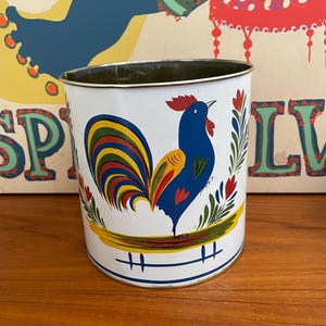 May include: White metal container with a colorful rooster design. The rooster is blue with a red comb and a yellow and red tail. The container has a green interior and is decorated with floral accents.