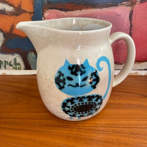 May include: A white ceramic pitcher with a blue and black cat design. The pitcher has a crackled glaze and a handle.