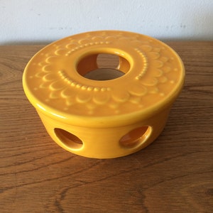 May include: A yellow ceramic tea light holder with a decorative floral design and three holes on the bottom.