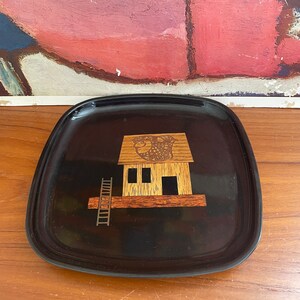 May include: A black, square-shaped tray with rounded corners. The tray features a wooden inlay design of a house with a ladder. The house has a wooden roof and is set against a black background.