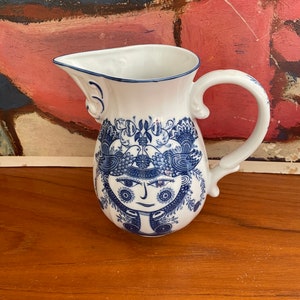 May include: A white ceramic pitcher with a blue and white floral design featuring a stylized face. The pitcher has a handle and a spout.