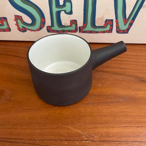 May include: A small, black ceramic pitcher with a white interior and a handle. The pitcher is sitting on a wooden surface.