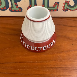 May include: A white and red ceramic wine bottle coaster with the word "VITICULTEURS" printed on the base. The coaster has a conical shape with a white, ribbed upper section and a red base.