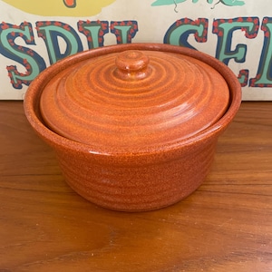May include: An orange ceramic casserole dish with a lid. The dish has a round shape with a slightly flared rim and a matching lid with a knob. The dish is sitting on a wooden surface.