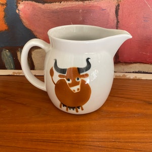 May include: White ceramic pitcher with a brown bull design. The bull has black horns and eyes. The pitcher has a handle and a spout.