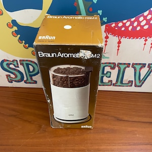 May include: A Braun Aromatic KSM2 coffee grinder in its original brown and white cardboard packaging. The box features the Braun logo, the product name, and a graphic showing the white cylindrical grinder filled with dark roasted coffee beans.