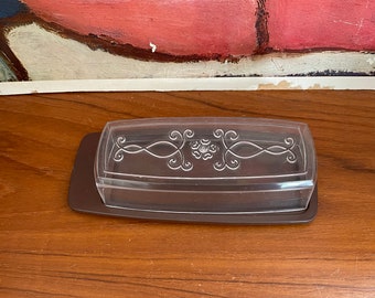 Könnte beinhalten: Eine transparente Butterdose aus Kunststoff mit braunem Sockel. Die Dose hat ein dekoratives Design auf dem Deckel.